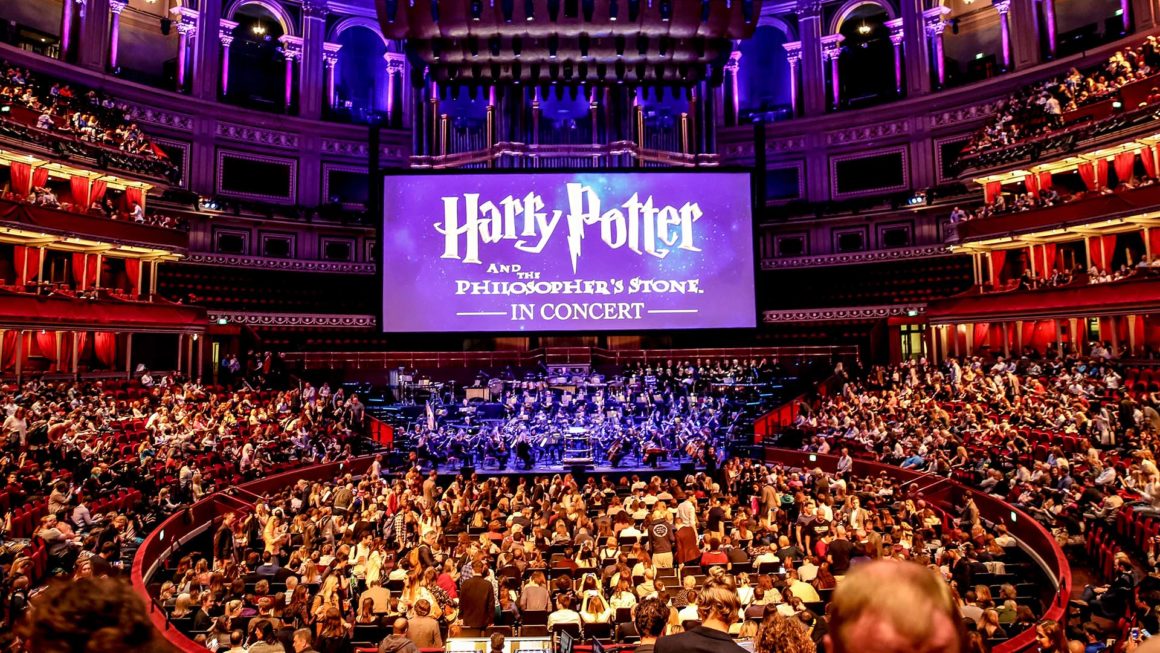 A packed concert hall with an orchestra playing beneath a large screen showing Harry Potter and the Philosopher's Stone in Concert