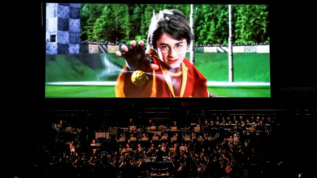 An orchestra performing live beneath a screen showing Harry Potter reaching for the Golden Snitch