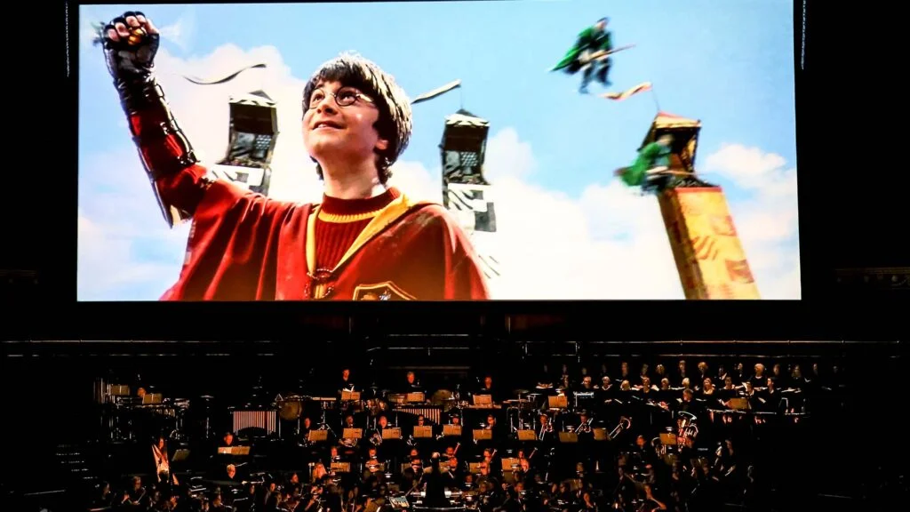 An orchestra performing live beneath a screen showing Harry Potter in Quidditch robes with a raised glove