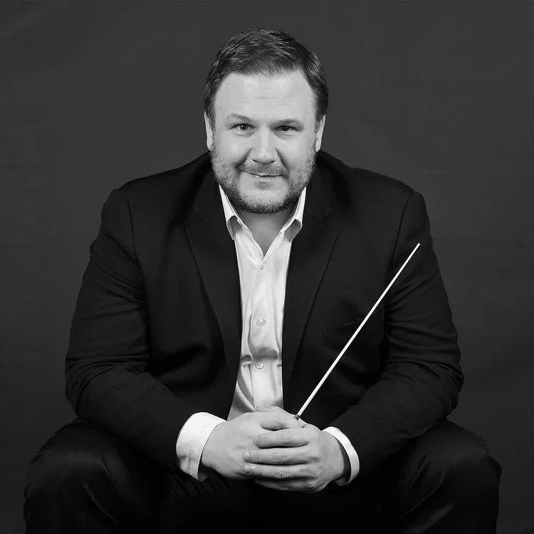 Black and white portrait of John Jesensky seated in a dark jacket, holding a conductor's baton