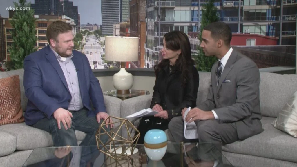 John Jesensky in a blue jacket on a studio sofa with two television hosts during an interview